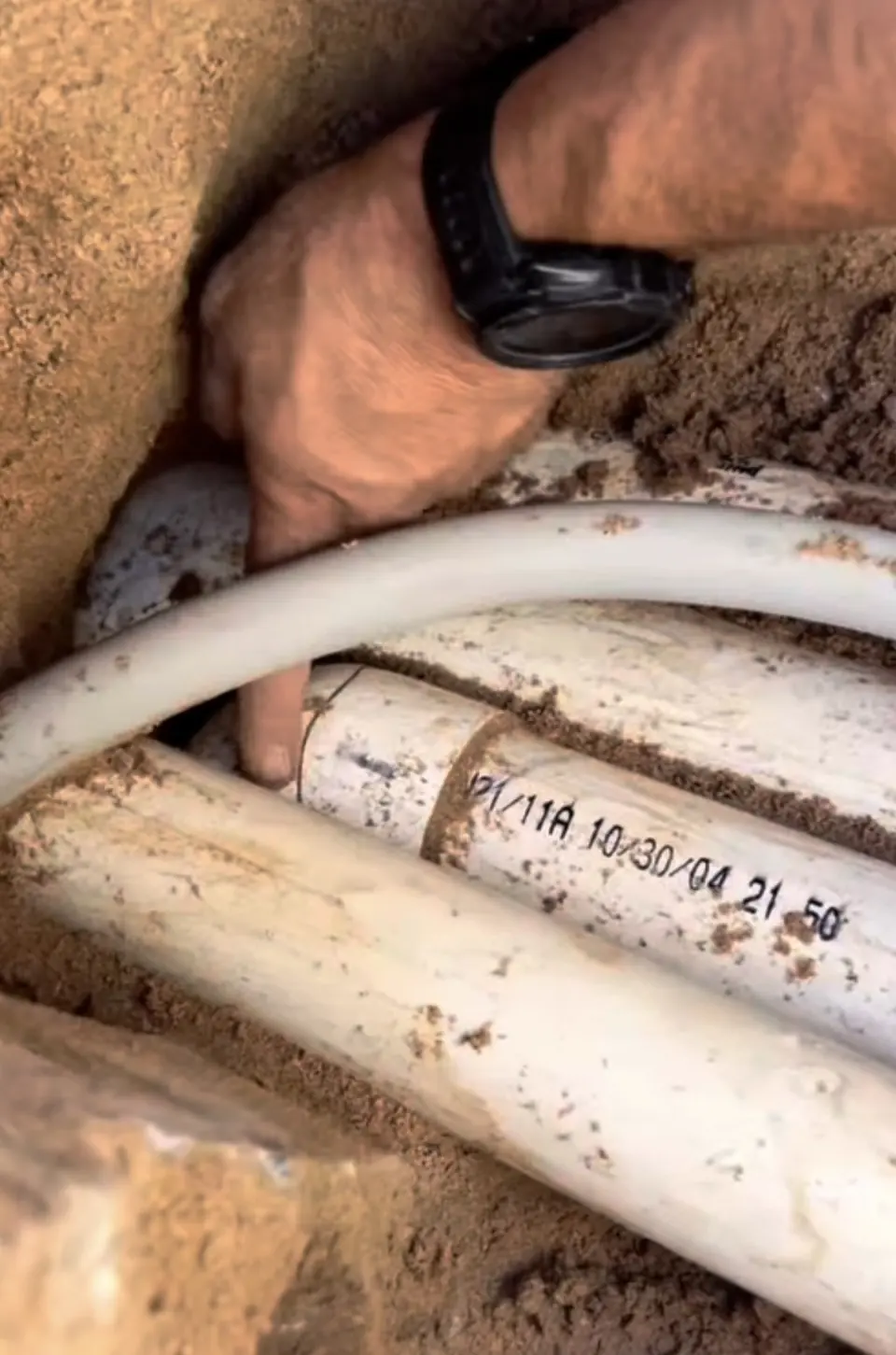 Hand pointing at white plastic pipes in a sandy trench